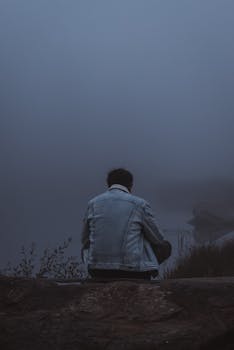 A person in a denim jacket sits alone in a misty, tranquil setting in Styria, Austria.