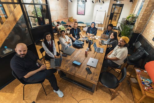 Group of colleagues gathered around a table in a modern office setting, collaborating on laptops.