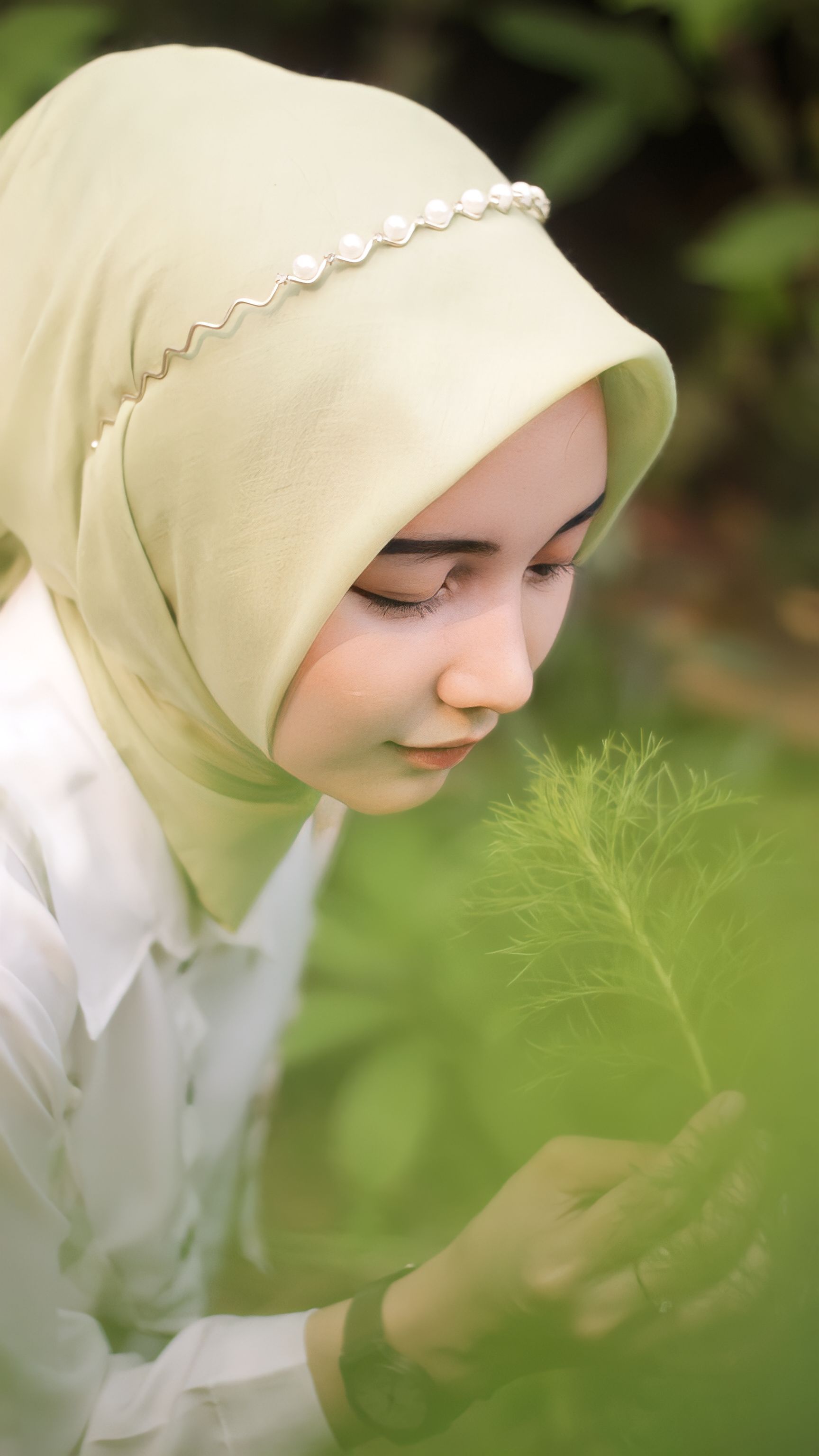 A serene portrait of a woman in a light green hijab holding a plant outdoors.