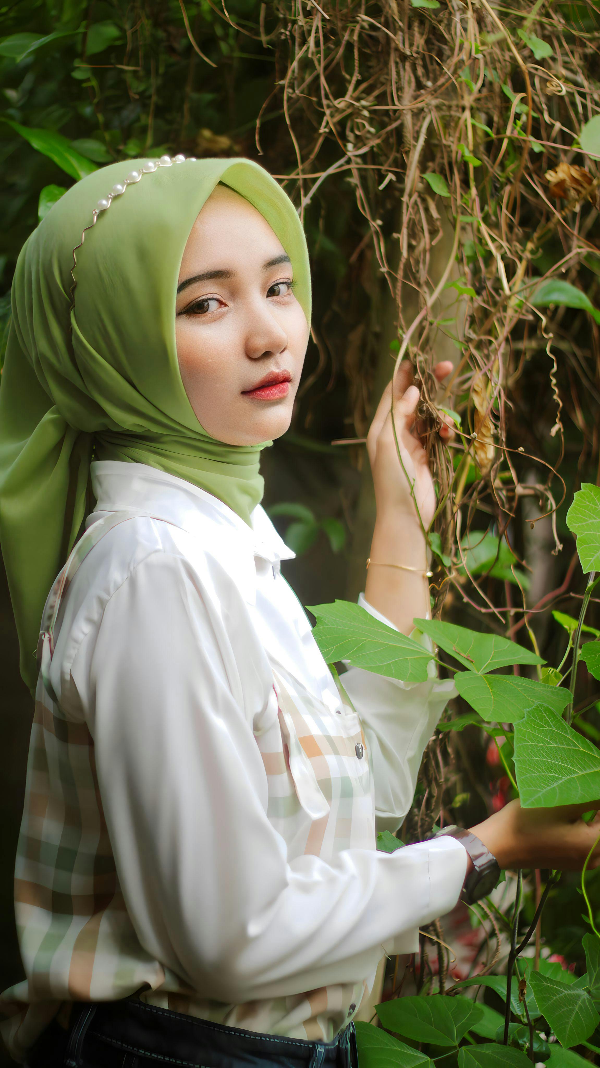 Portrait of a woman in a green hijab surrounded by lush greenery outdoors.