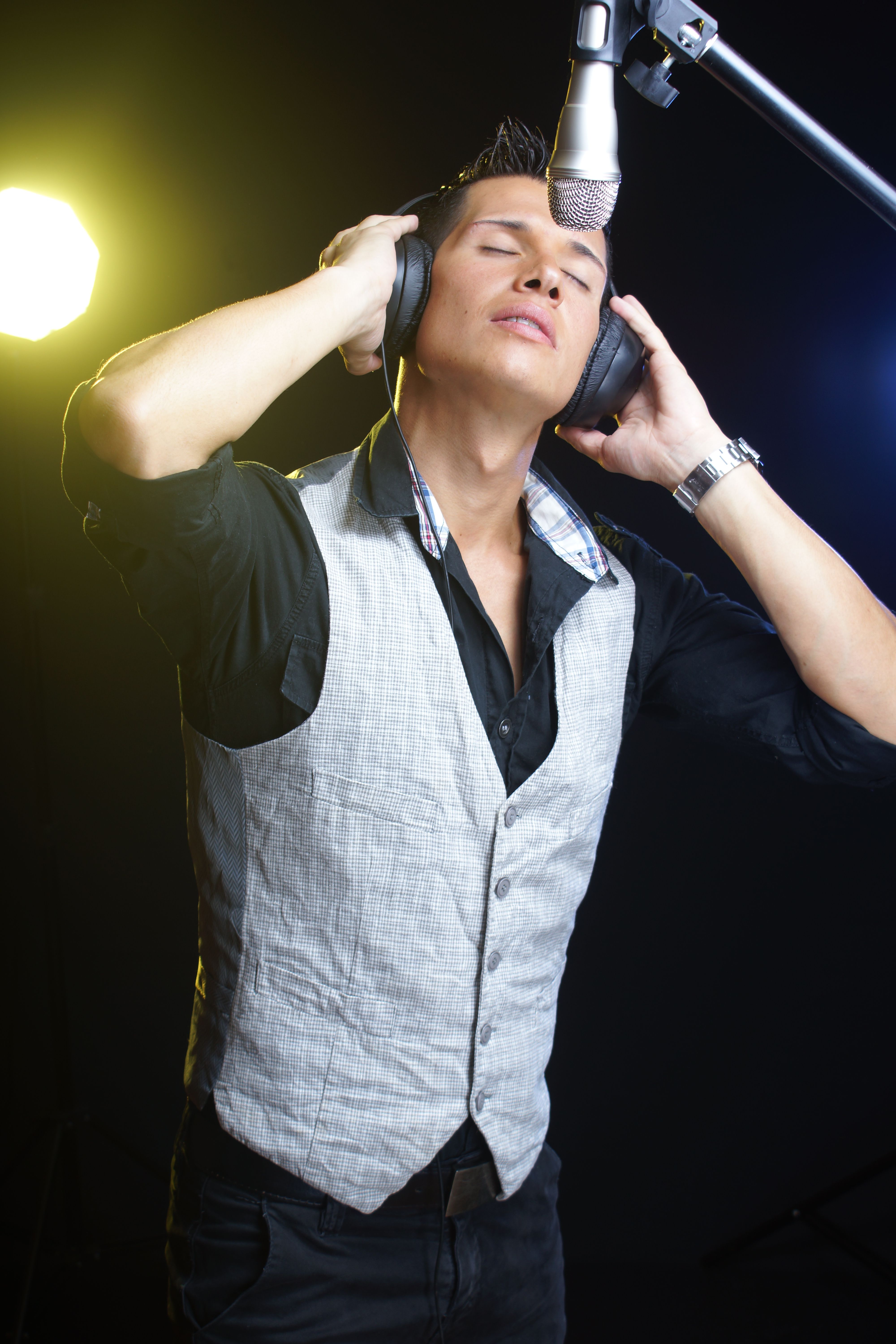 A young male singer in a studio wearing headphones and singing into a microphone.