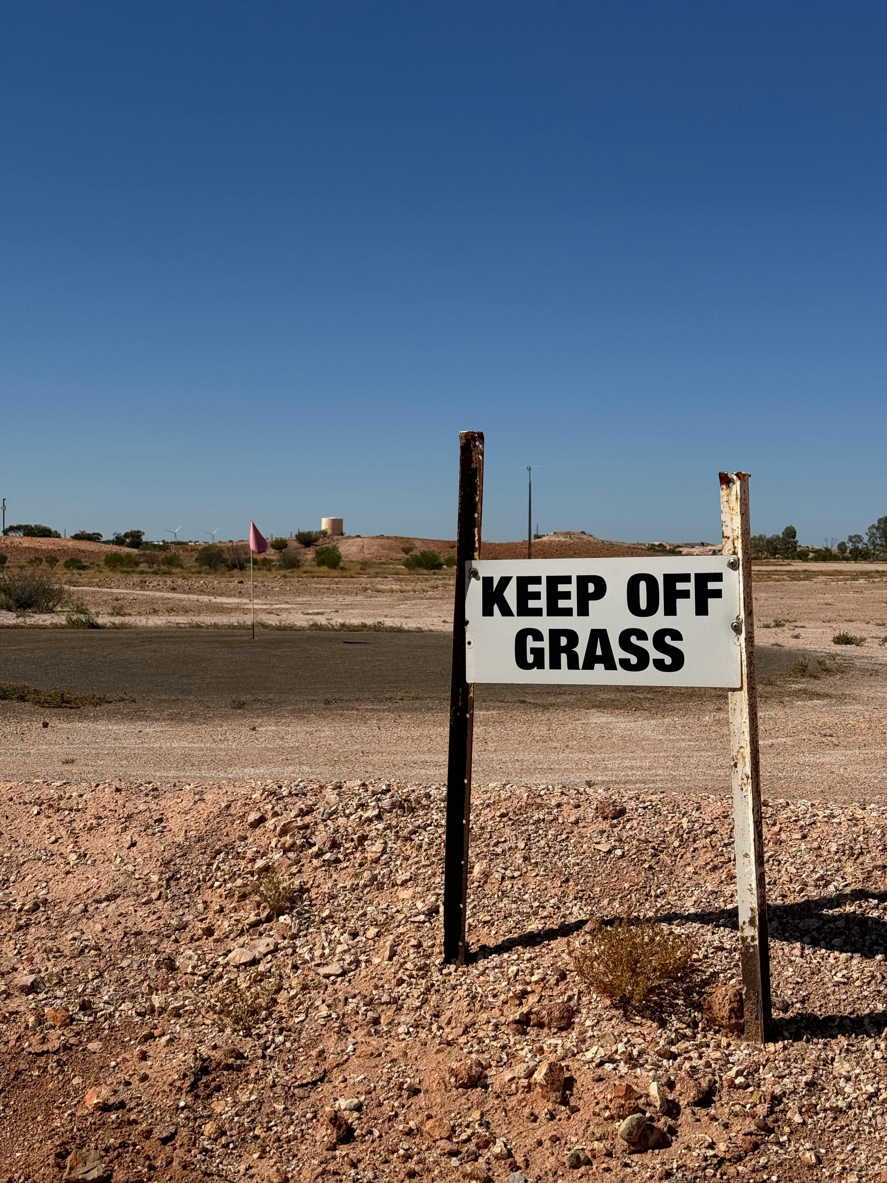 A dry, barren desert area with a 'Keep Off Grass' sign under clear skies.