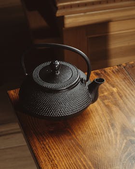 A traditional black cast iron teapot sits elegantly on a sunlit wooden table indoors.