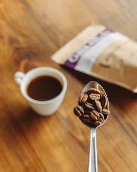 A cup of coffee with coffee beans on a spoon, blurred coffee bag in background.