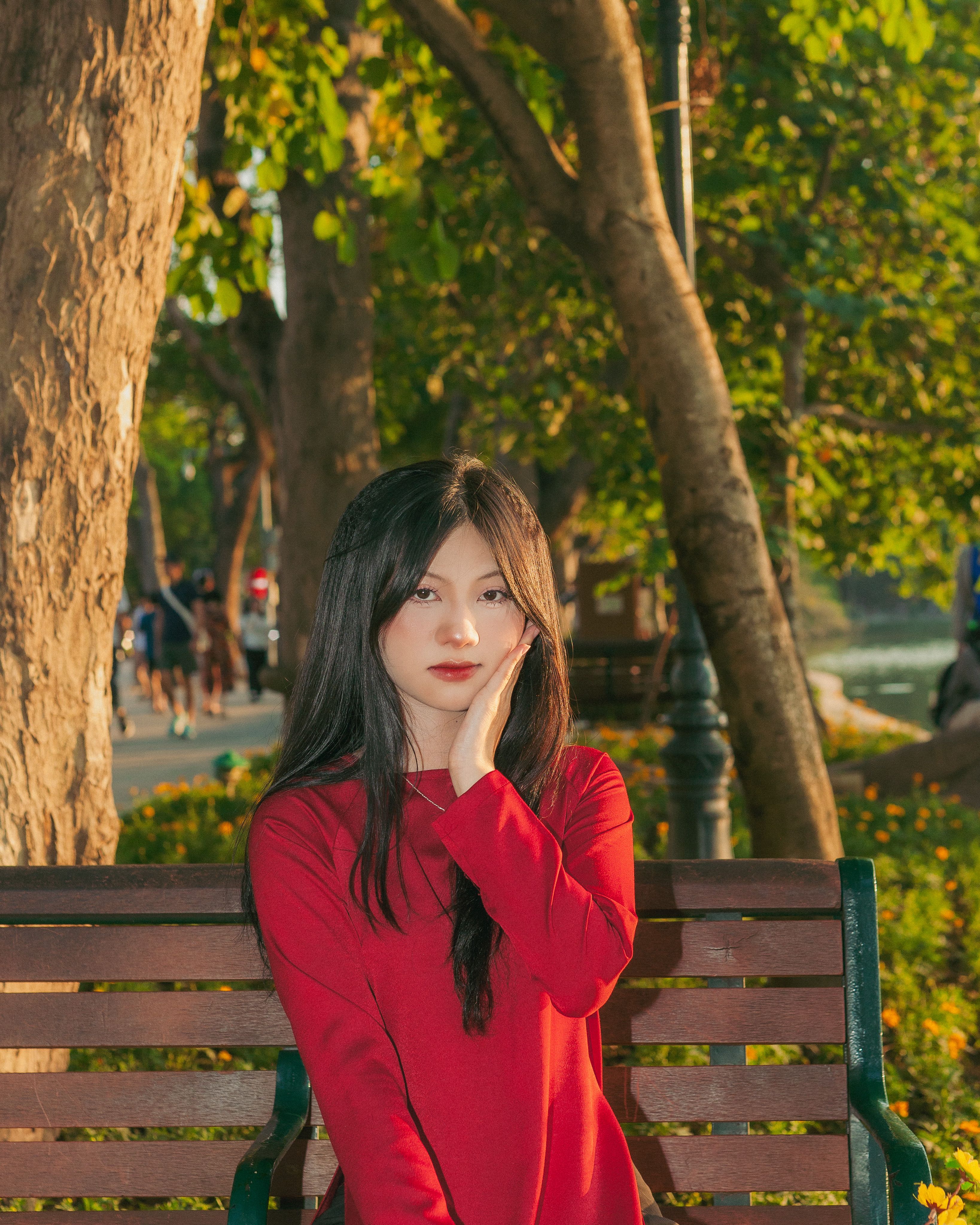 Portrait of a woman in a red ao dai at Hoan Kiem Lake, Hanoi, surrounded by lush greenery.
