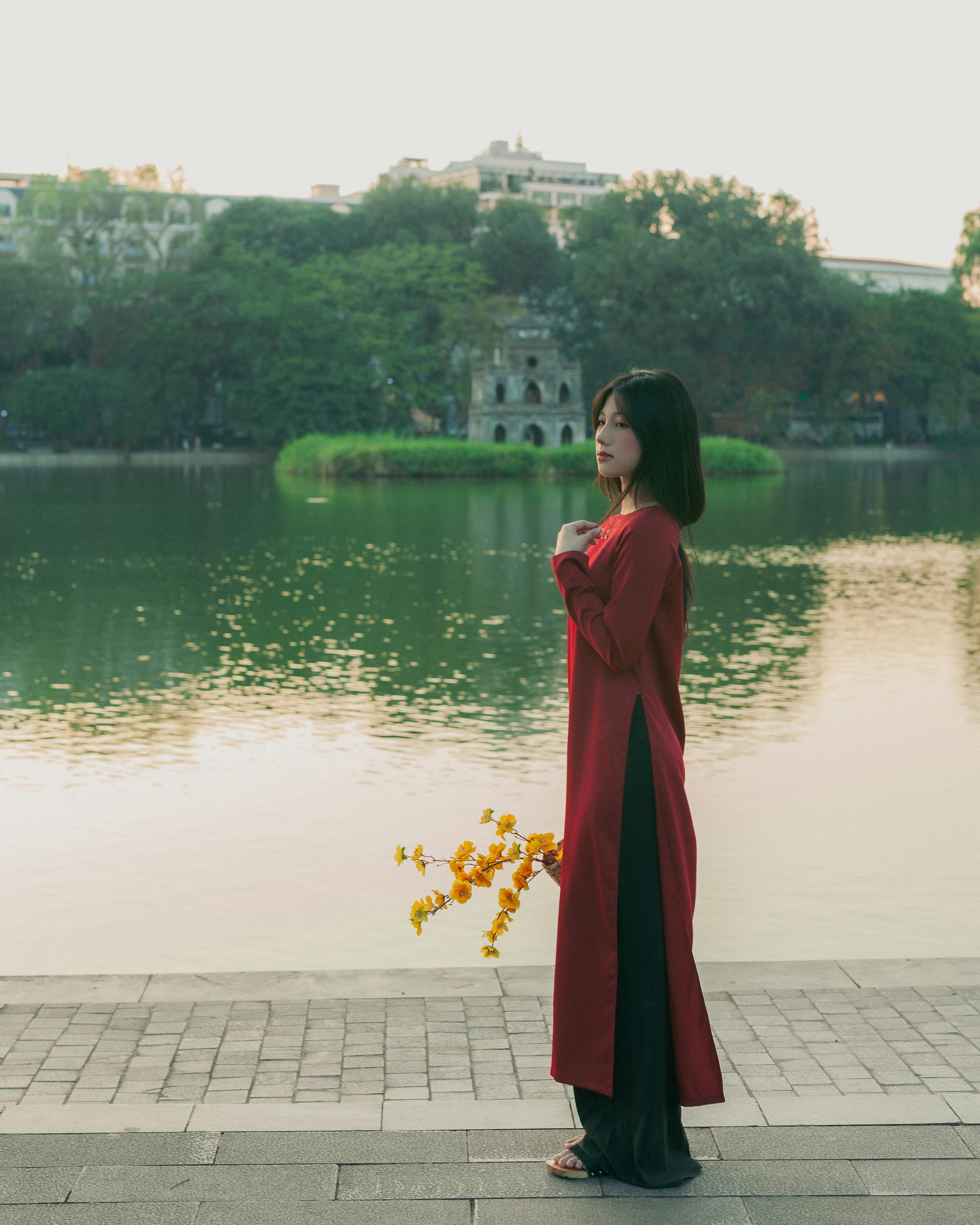 A woman in traditional red ao dai stands by Hoan Kiem Lake in Hanoi during a peaceful sunset.