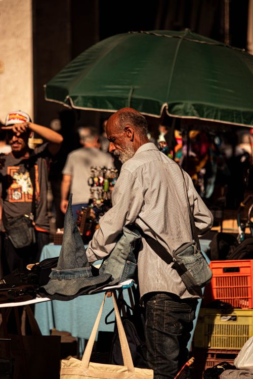 Free Elderly vendor managing a stall under a green umbrella in a bustling street market. Stock Photo