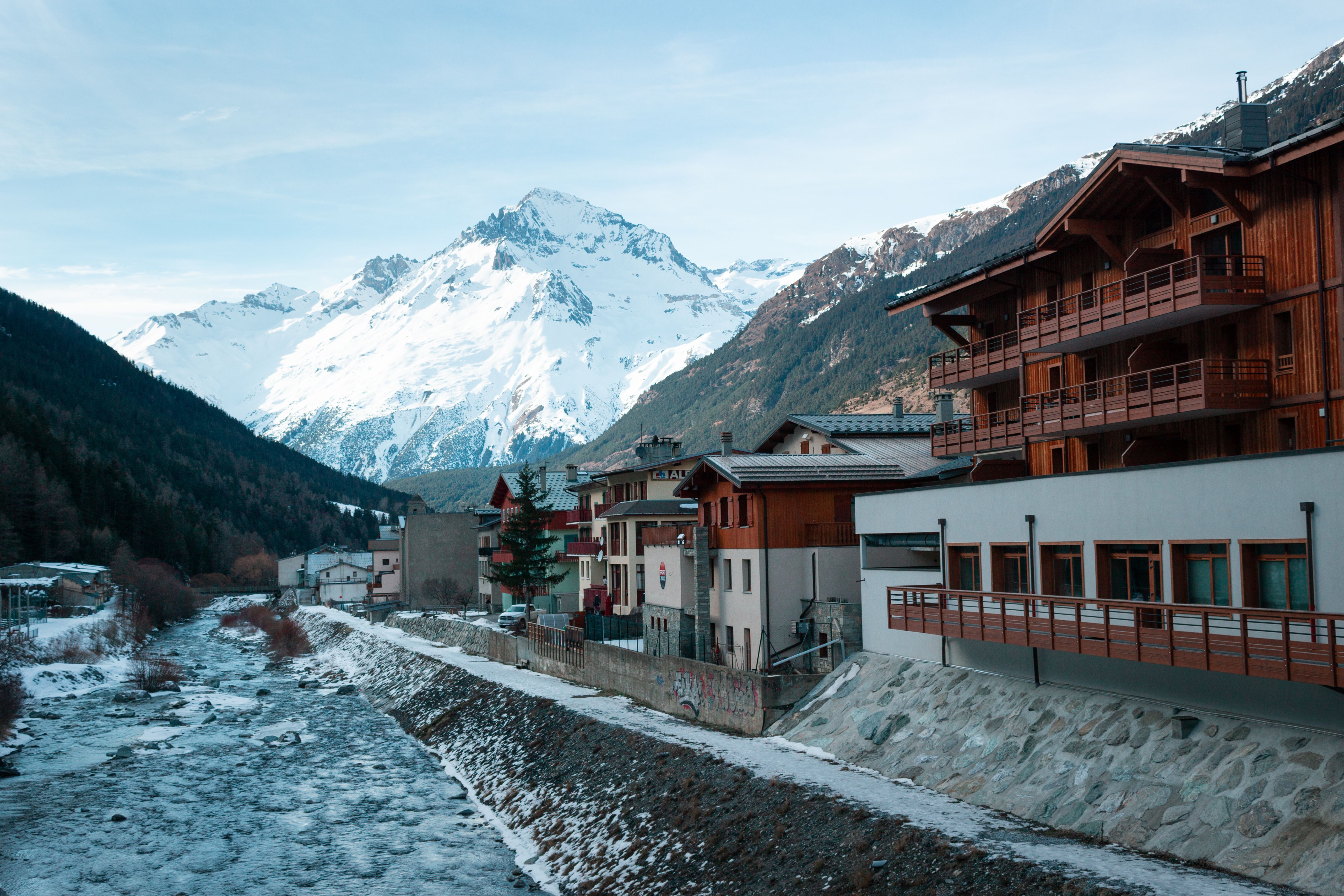 A valley-floor alpine village beside a river in early winter, timber balconies above a road that runs up to the front doors