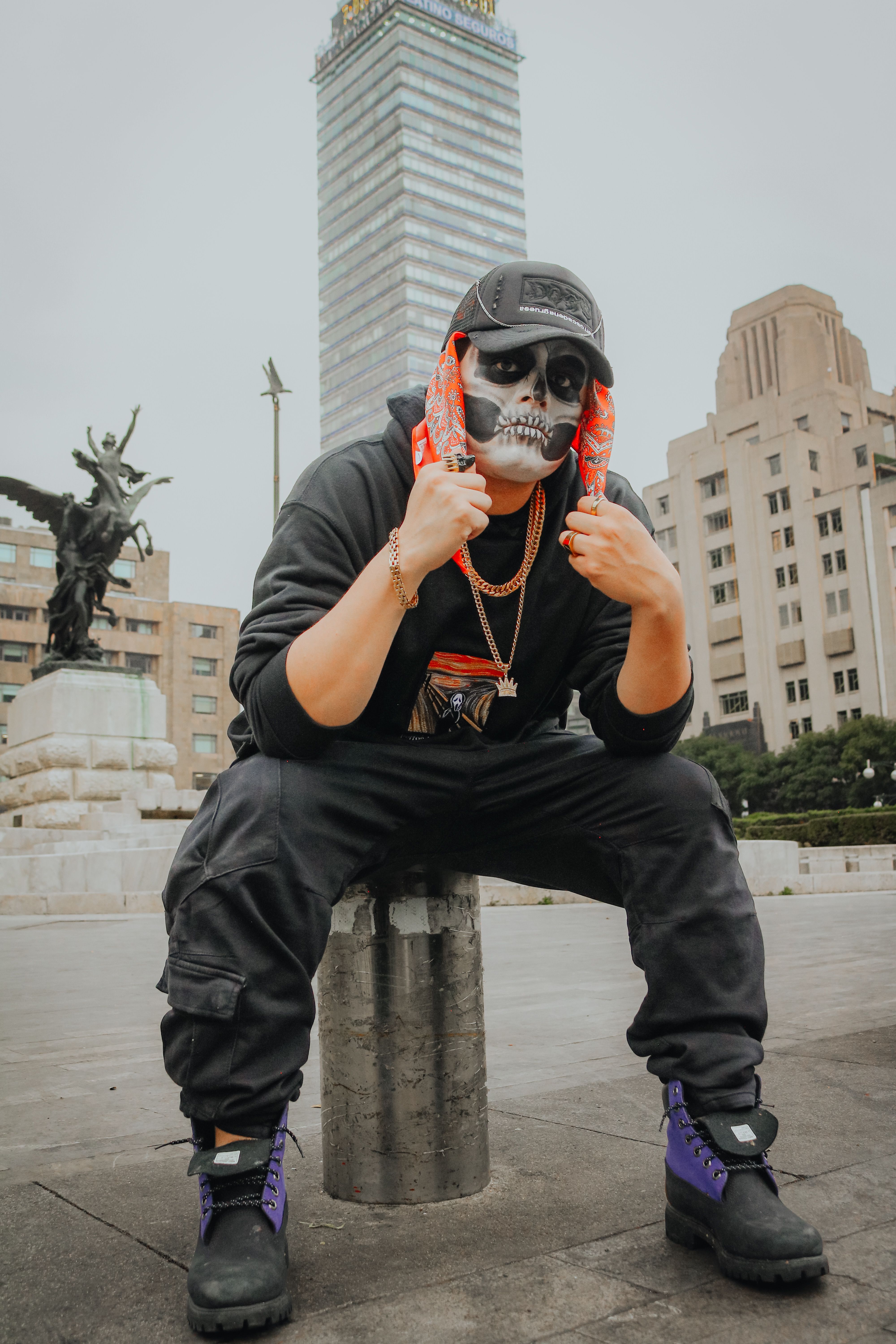 Person wearing a mask seated in a plaza with a tall skyscraper and statue in the background.