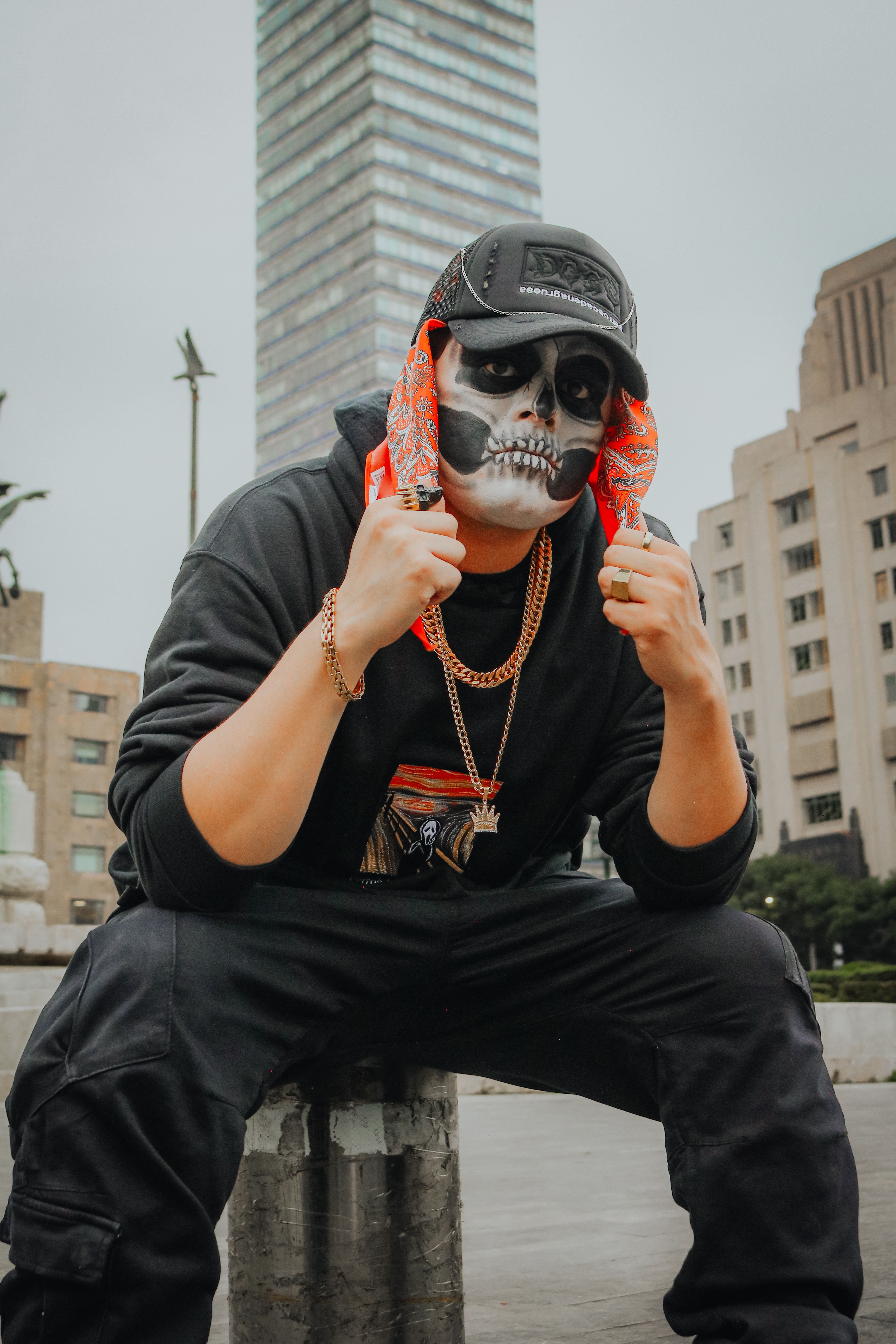 Person wearing a skull mask and bandana in an urban setting with skyscraper backdrop.