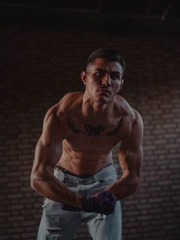 Shirtless muscular man with tattoos flexing against a brick wall indoors.