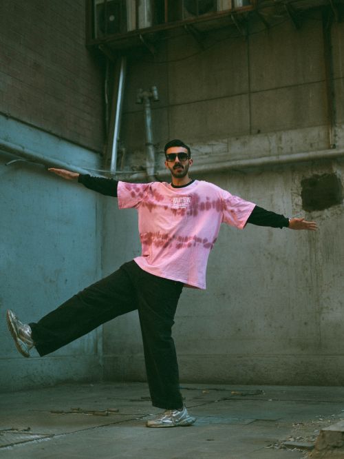 Free Stylish man balances playfully in an urban alley, wearing a pink tie-dye shirt and sunglasses. Stock Photo