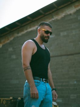 Stylish man in sunglasses and tank top stands confidently against an urban backdrop.