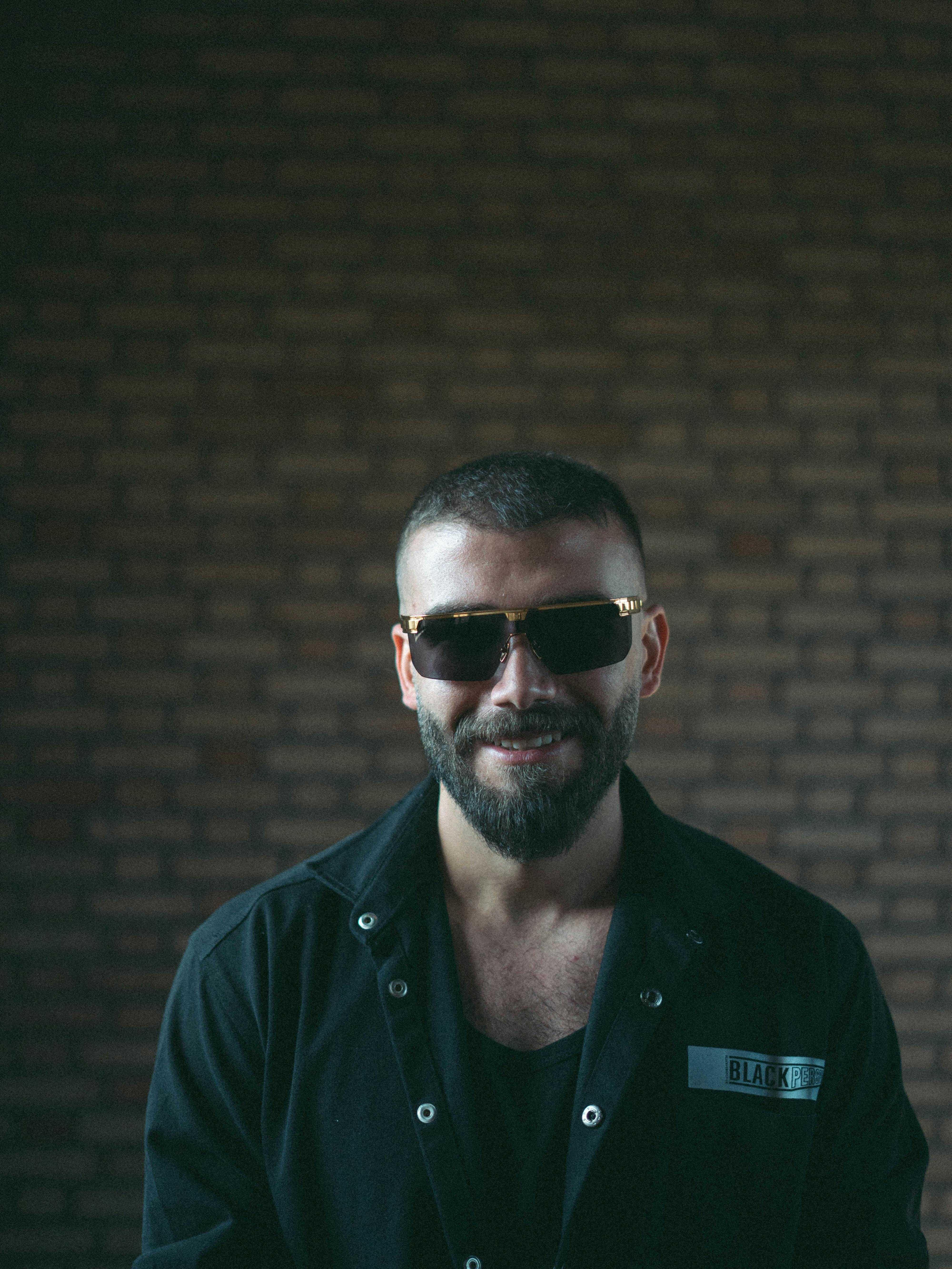 Confident man wearing sunglasses with a stylish beard against a brick backdrop, exuding modern fashion vibes.
