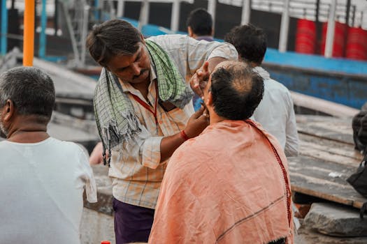 A street barber provides a close shave to a client in a bustling outdoor market setting.