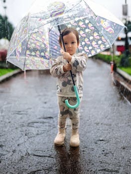 A young child stands under a cartoon-themed umbrella during a light rain on a park path.