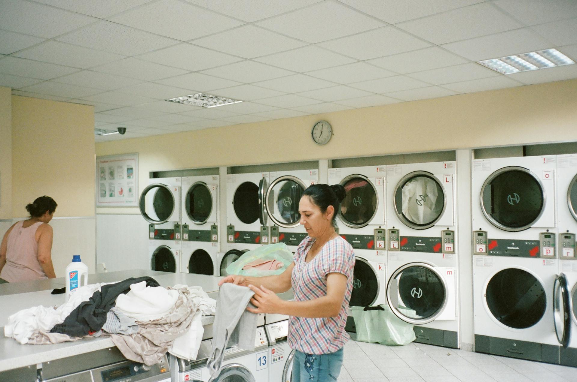 A woman folding clothes in a modern laundromat with various washing machines in the background.