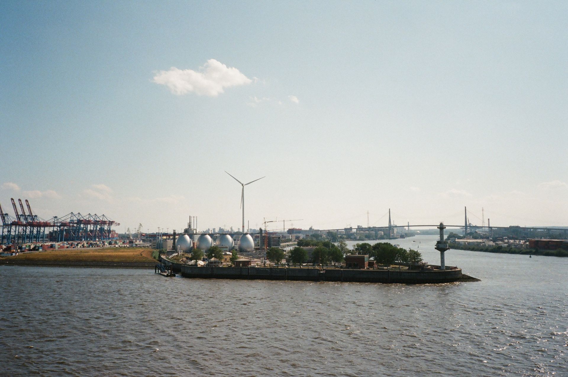A scenic view of an industrial area by a river, featuring a wind turbine and bridges under a blue sky.