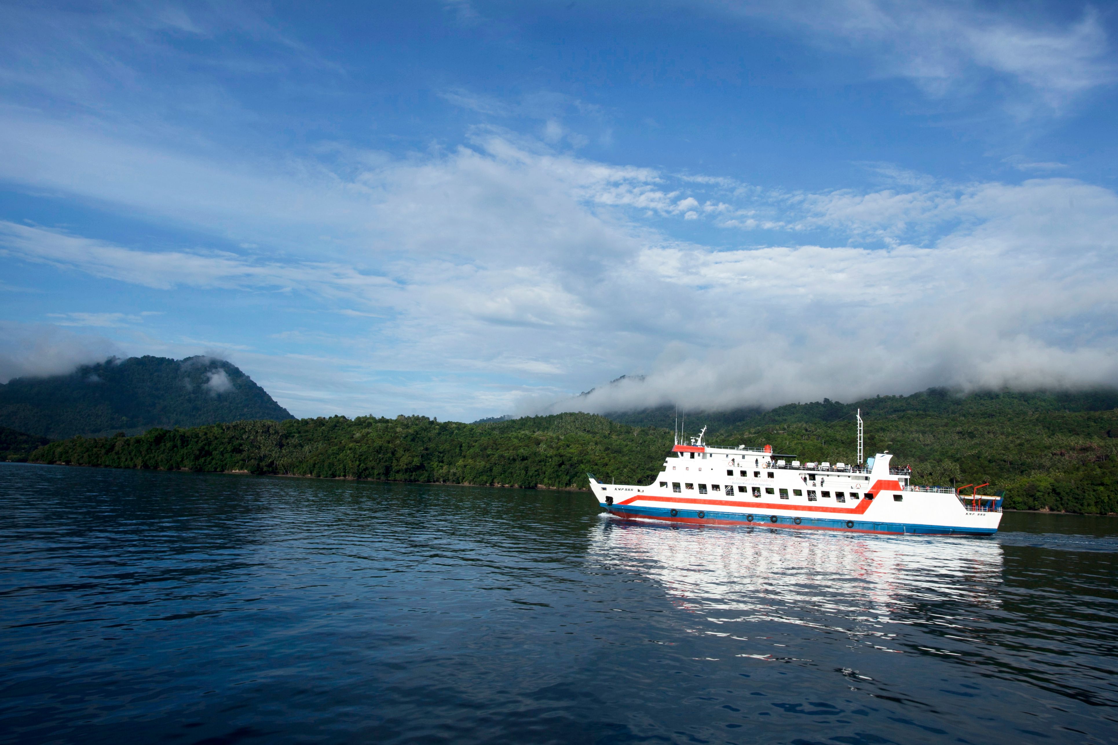 A ferry sails on a calm lake against a backdrop of lush, green hills under a cloudy sky.