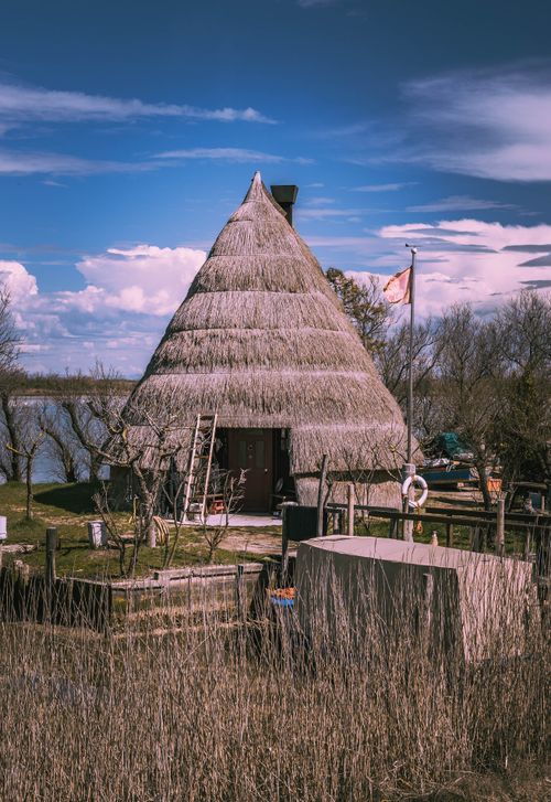 Free A rustic thatched hut by the riverside surrounded by lush spring foliage under a bright blue sky. Stock Photo