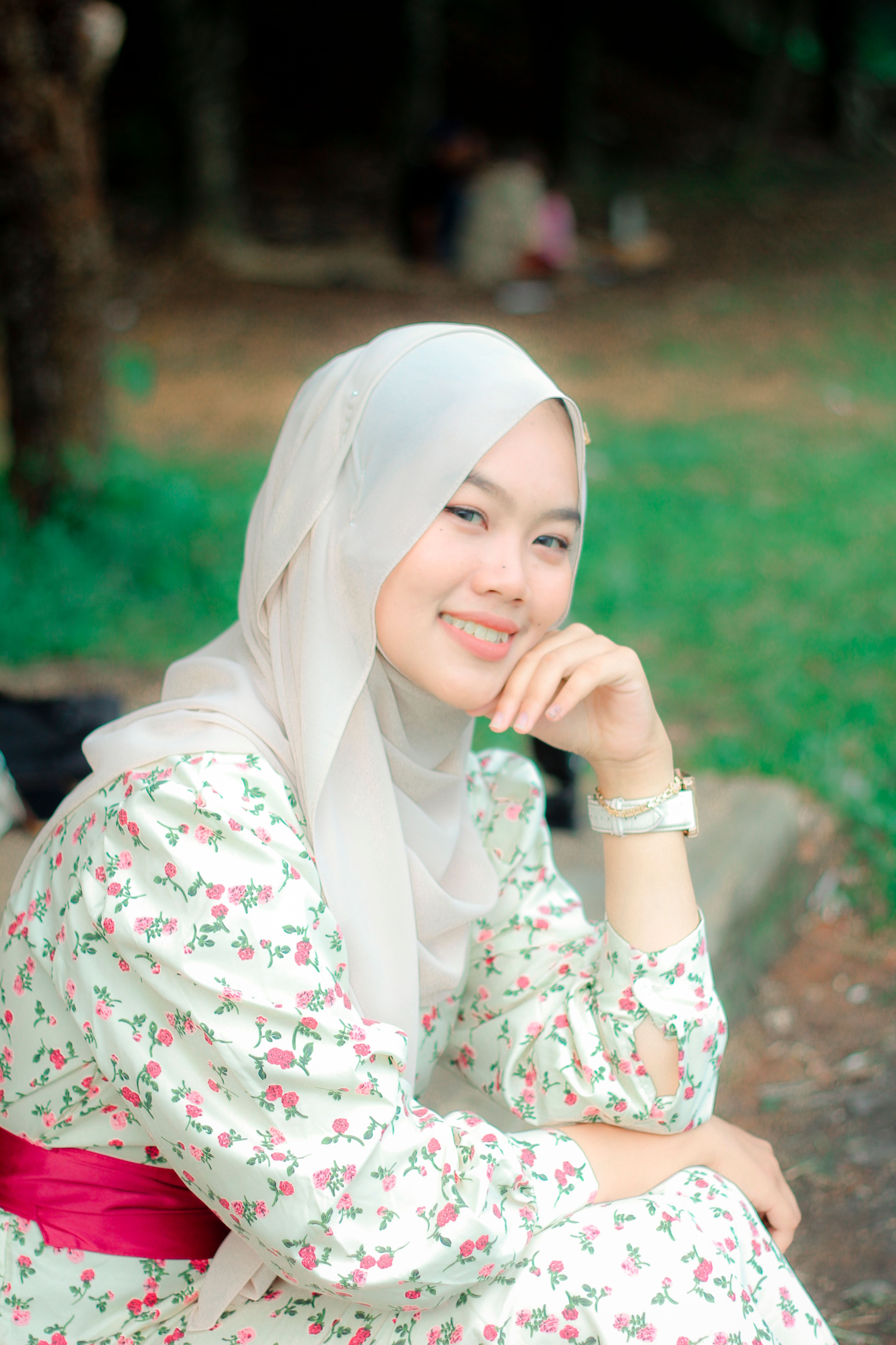 Portrait of a smiling woman wearing a hijab, sitting in a park with greenery.