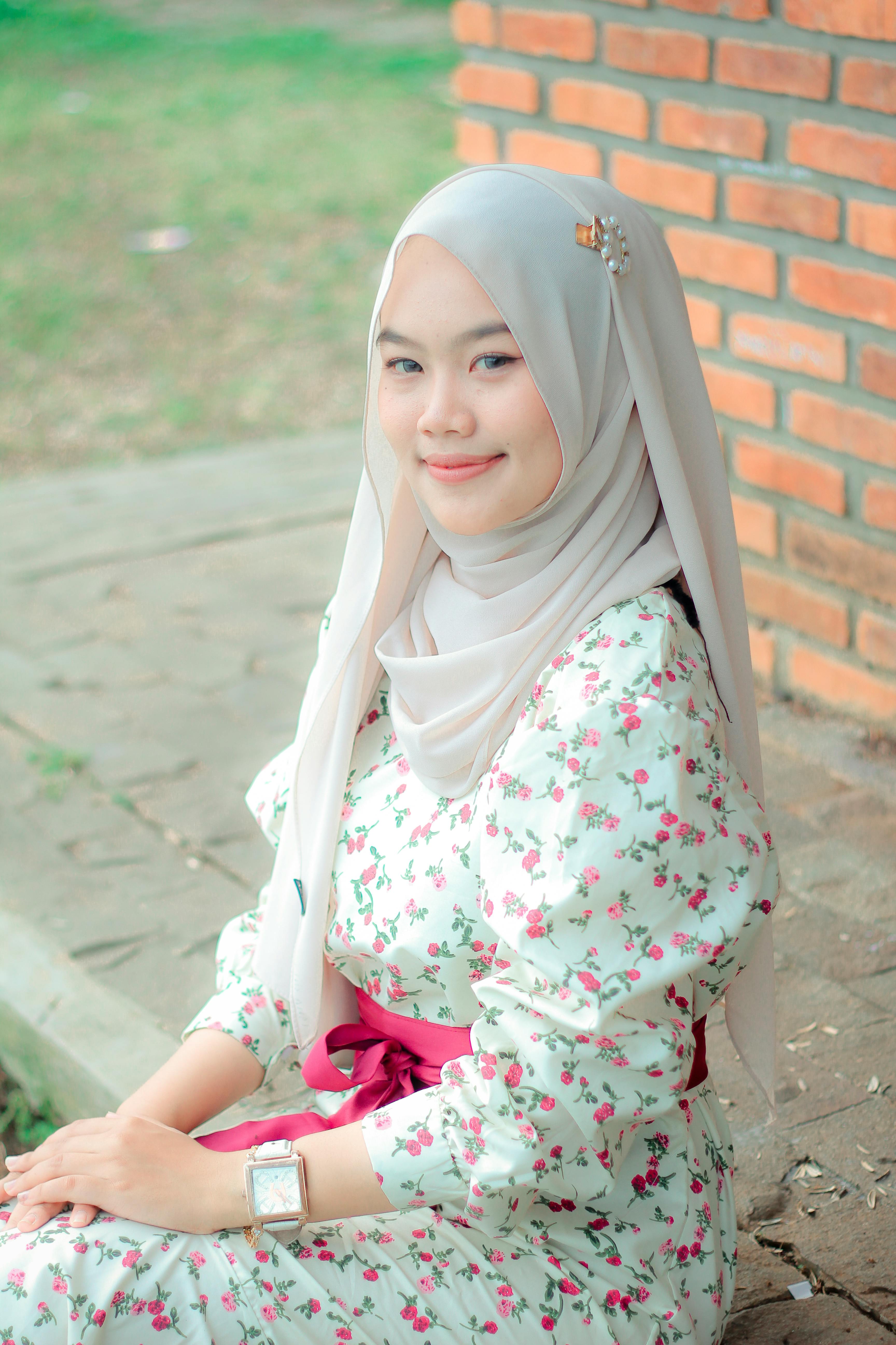A cheerful woman in a floral dress and hijab sits outdoors, exuding warmth.