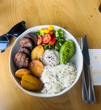 Colorful bowl with rice, avocado, patties, and veggies on wooden table.