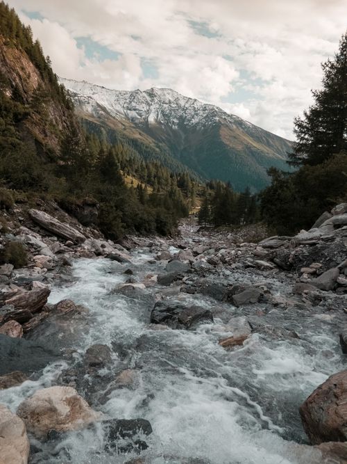 Free A serene mountain river flowing through lush forests with a stunning backdrop of snow-capped peaks. Stock Photo