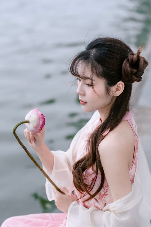 Free A woman in traditional attire peacefully holding a lotus flower by a calm waterfront. Stock Photo