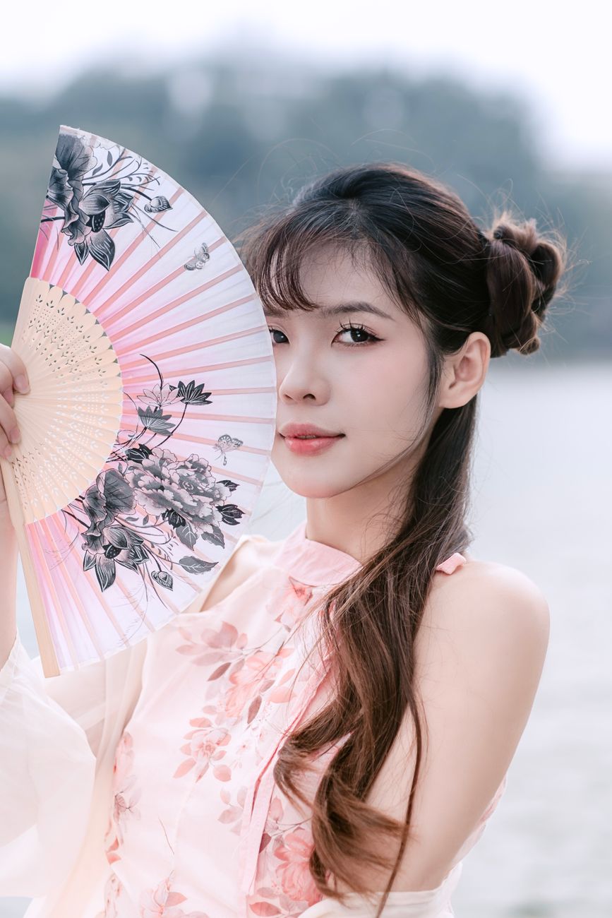 A young woman in a floral dress holds a decorative fan, exuding elegance and grace outdoors.
