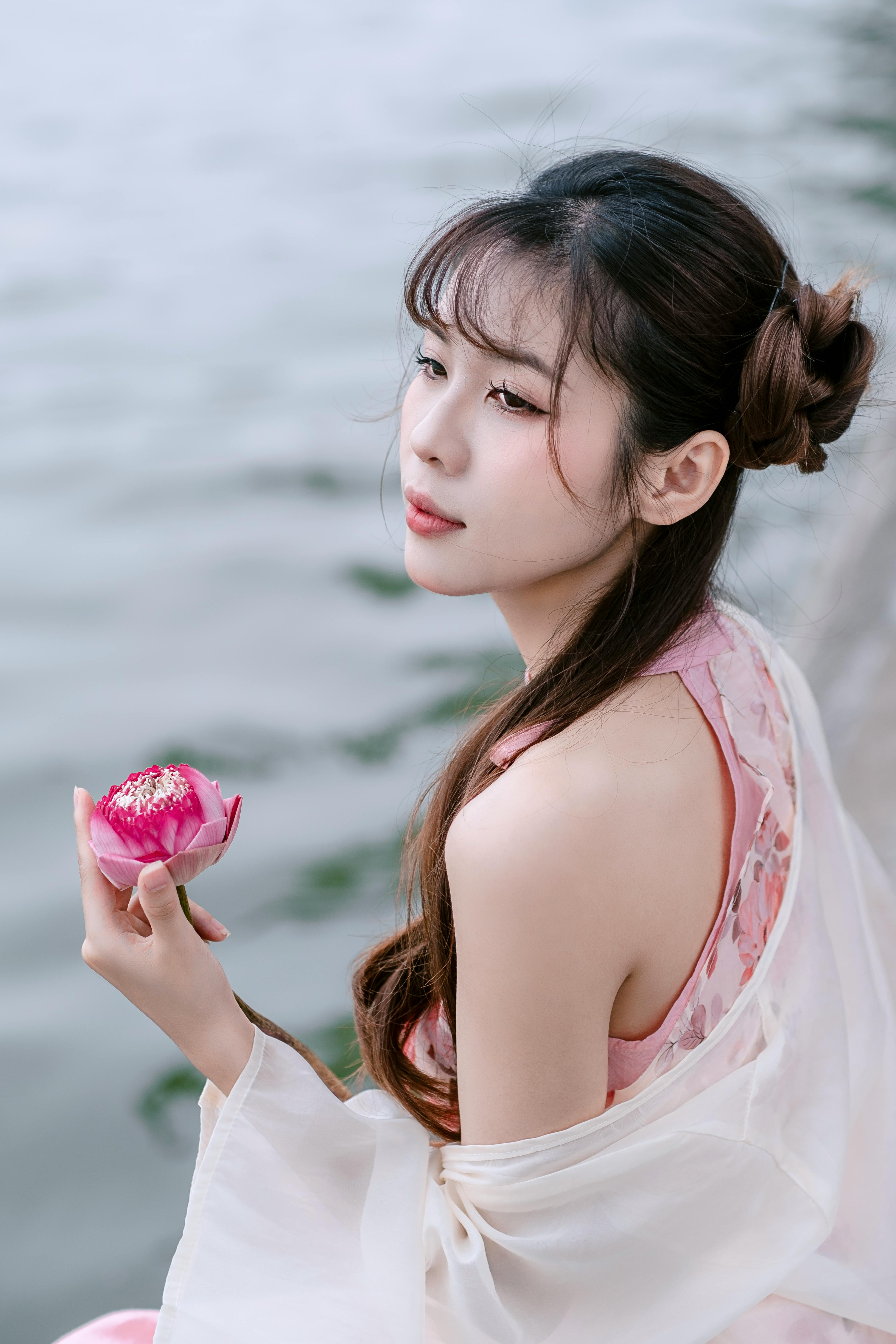 Elegant young woman holding a lotus flower by the water's edge, wearing traditional attire.