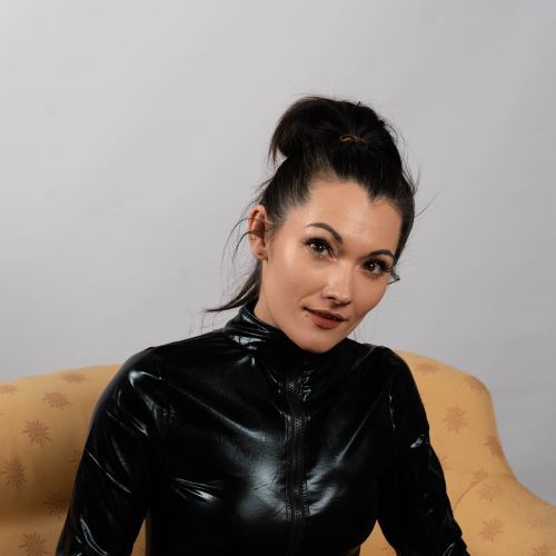 Free Elegant woman in black shiny outfit seated indoors, exuding confidence. Stock Photo