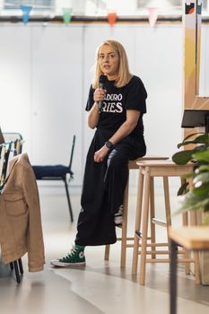 Woman sitting on a stool speaking into a microphone at an informal gathering.