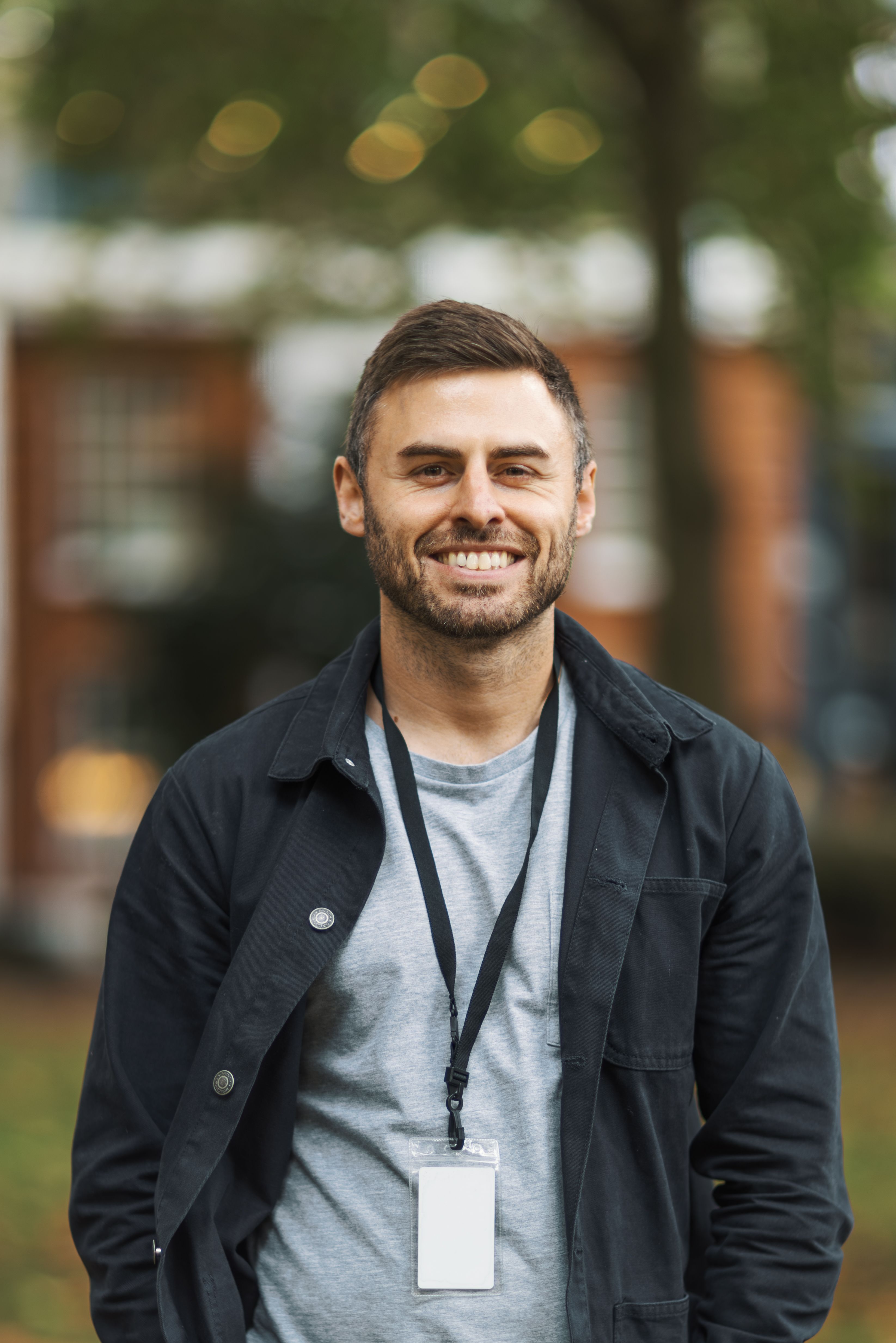 Portrait of a smiling man in casual attire with a lanyard, standing outdoors.