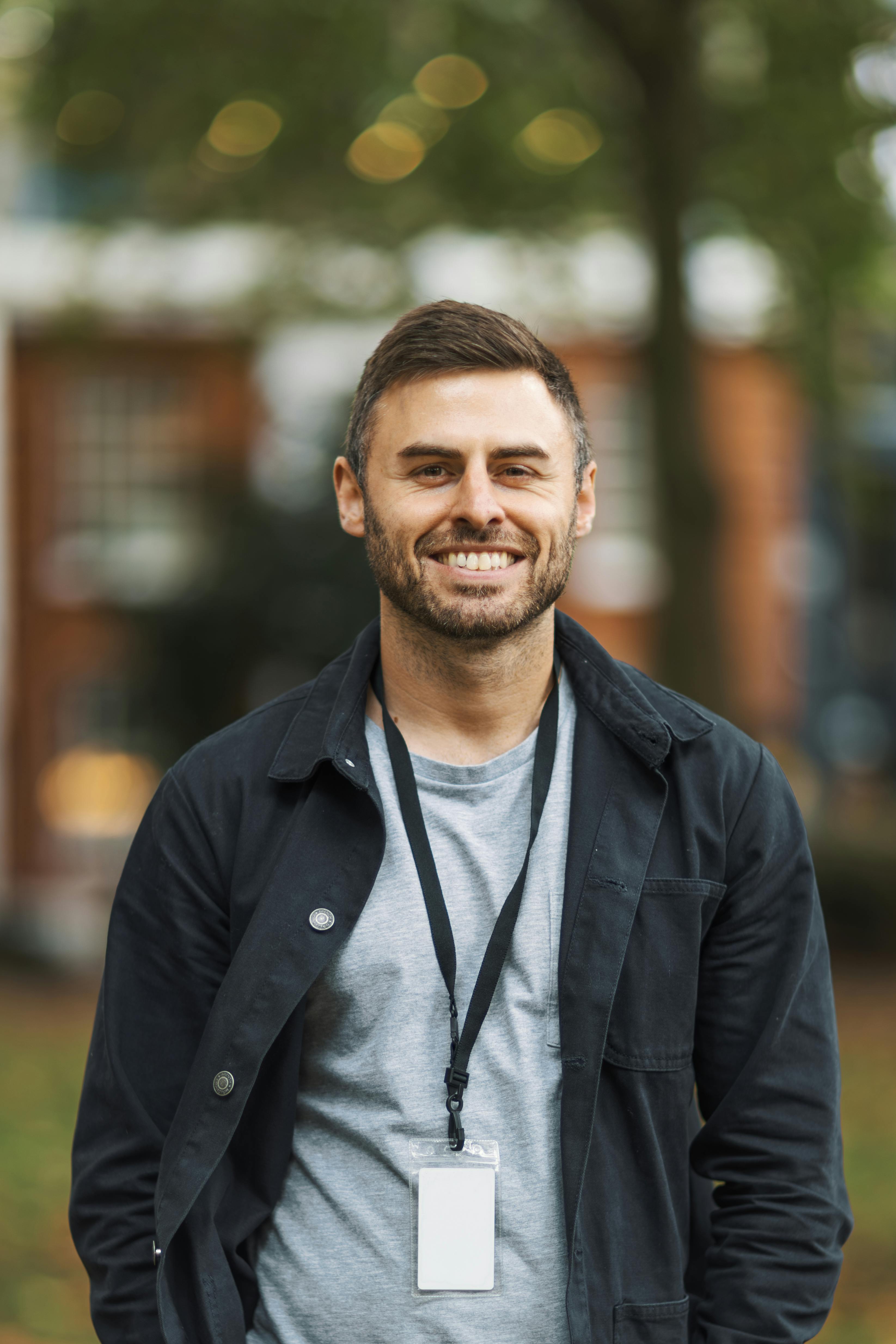 Ilmainen Muotokuva hymyilevästä miehestä rennossa asussa ja kaulanauhalla seisovasta ulkona. Kuvapankkikuva