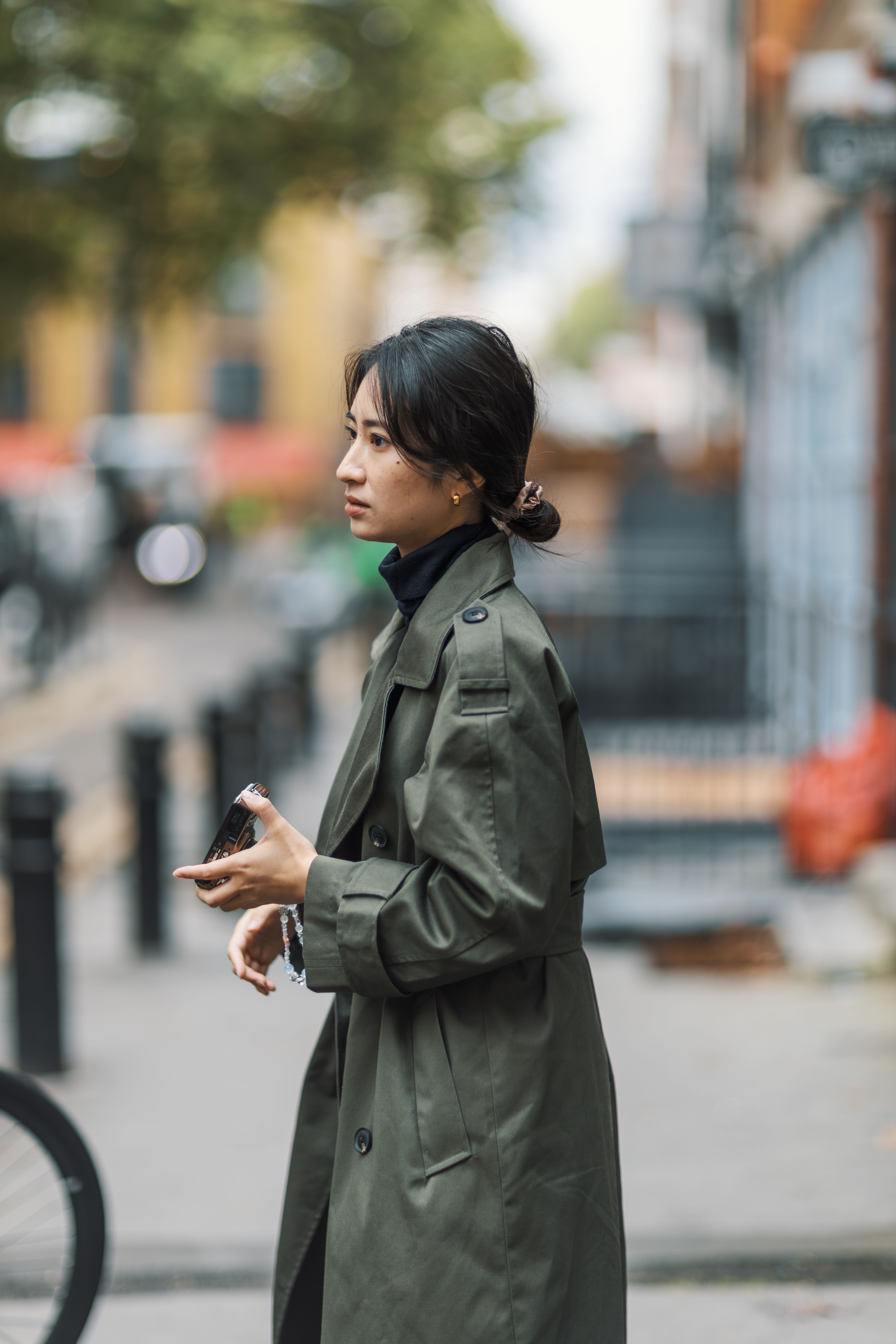 A fashionable woman in a green trench coat walking through a city street holding a smartphone.