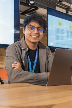 Young adult male working on a laptop in a modern office setting, smiling at the camera.