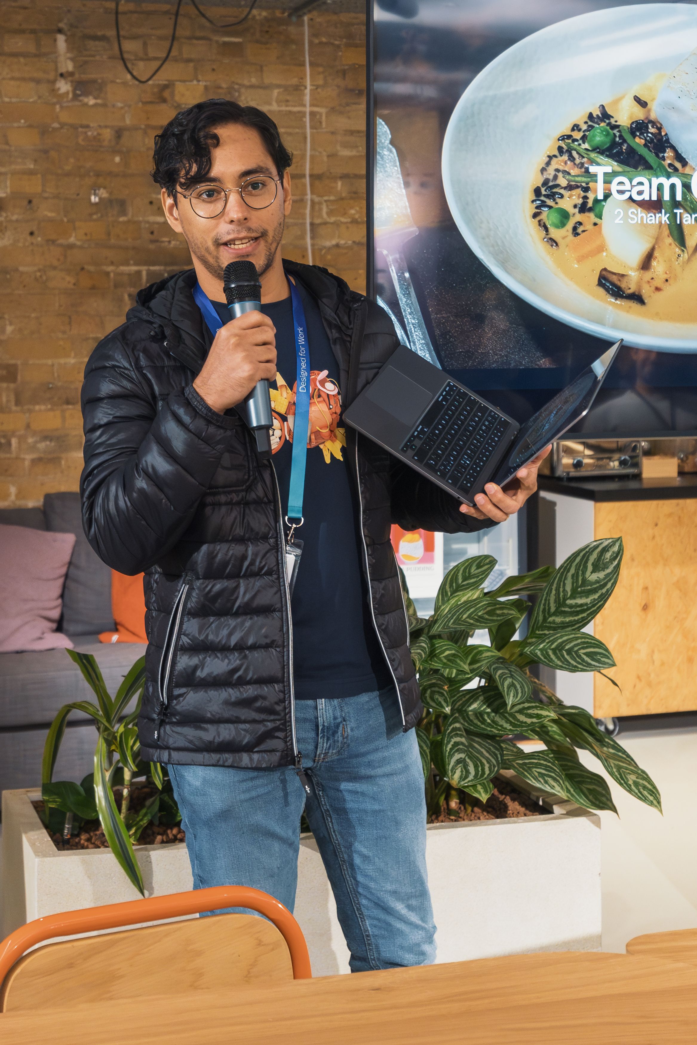 A young man presents using a laptop and microphone in a modern office setting.
