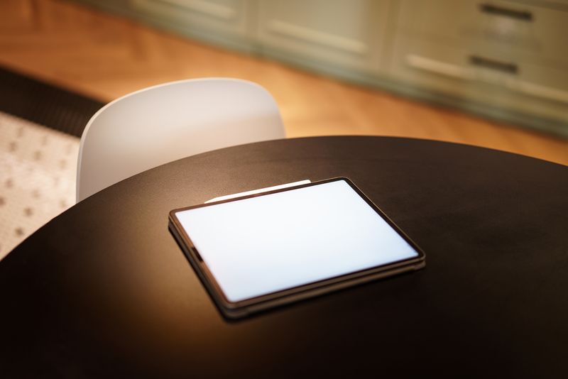 A tablet with a blank screen resting on a sleek black round table in a warm, modern kitchen setting.