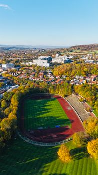 Aerial shot of a sports stadium amidst fall foliage with scenic city and hills in the backdrop.