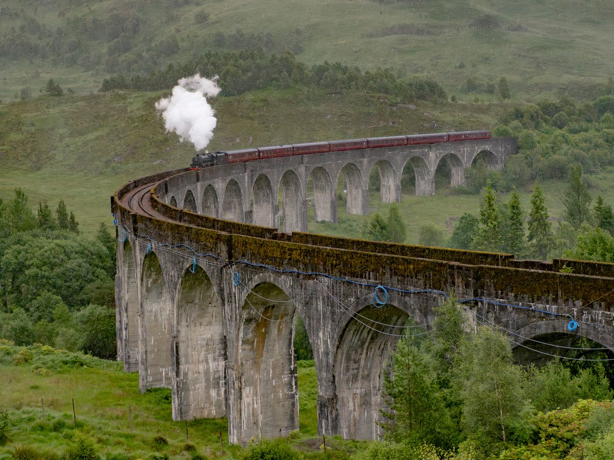 Tren cu aburi traversând viaductul Glenfinnan din Highlands, cu dealuri verzi în jur