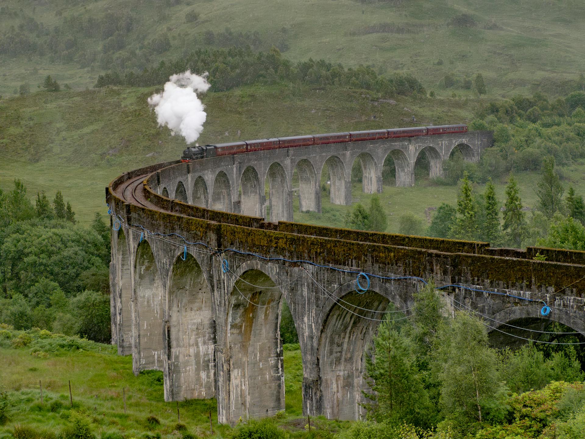 Steam train crosses the iconic Glenfinnan Viaduct in lush Scottish landscape.