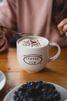 A person enjoys a frothy cappuccino in a 'Coffee Cup' mug at a cozy cafe.