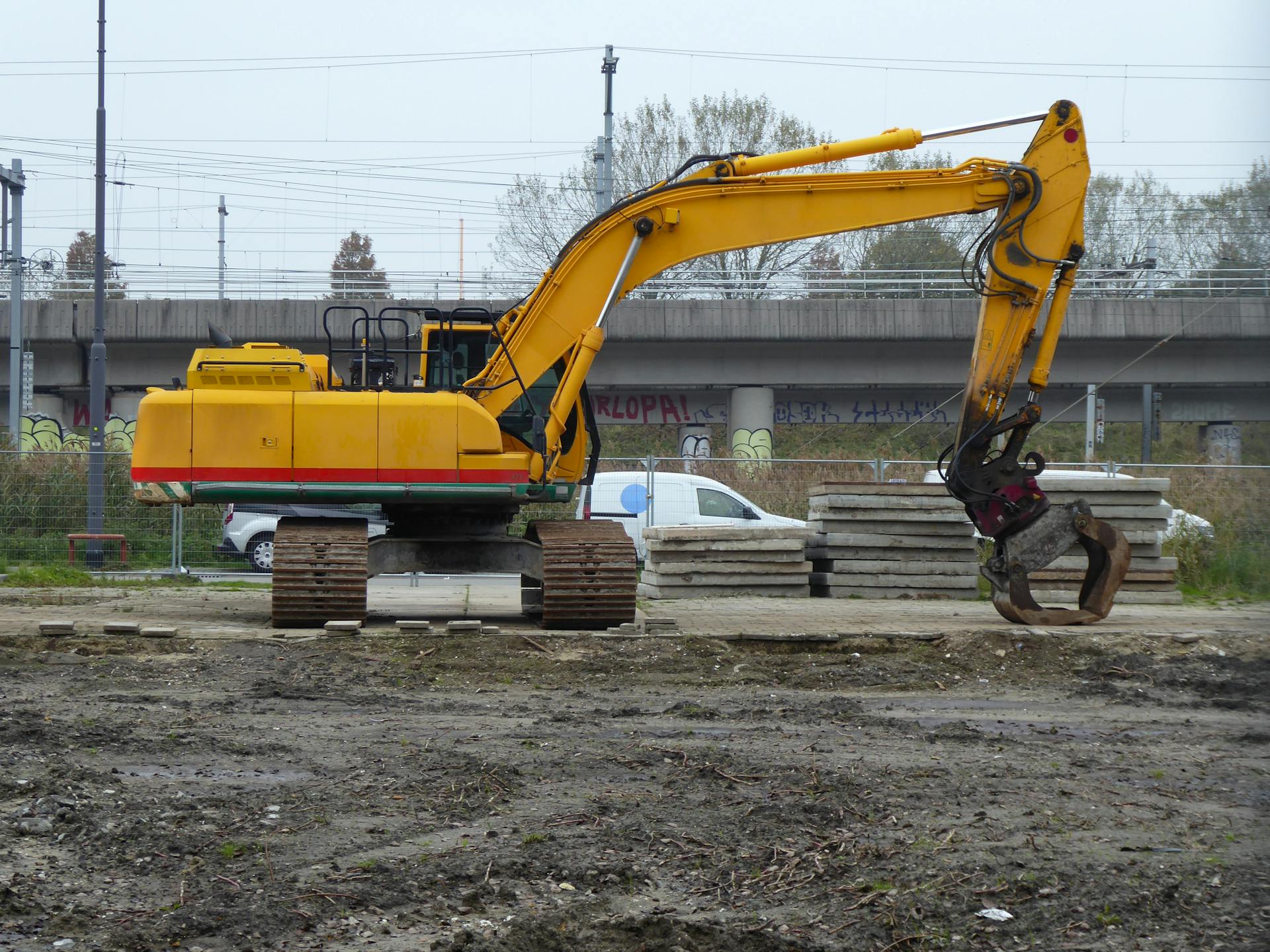 Yellow excavator at a construction site in Amsterdam, with concrete blocks and railway in the background.