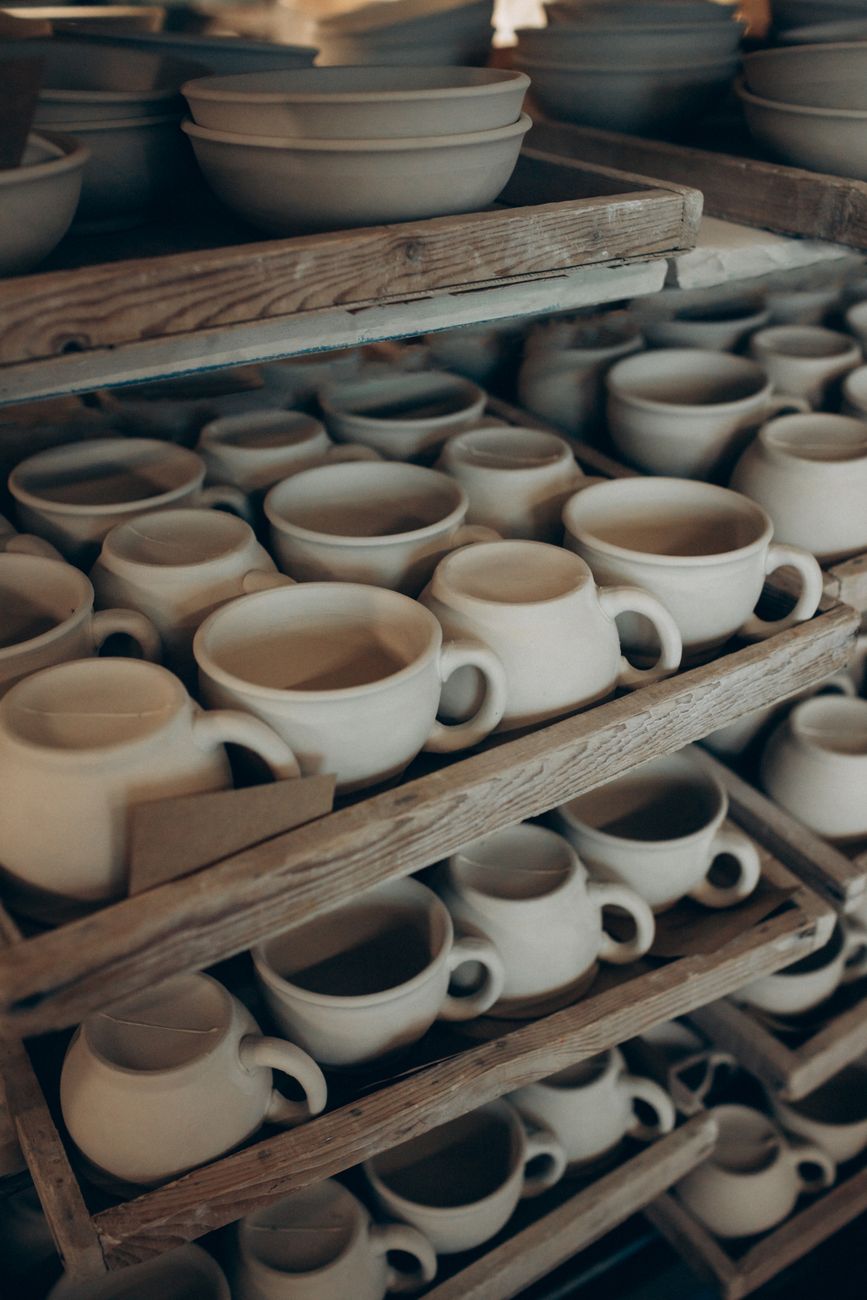Rows of handmade ceramic cups and bowls on rustic wooden shelves in a pottery studio.