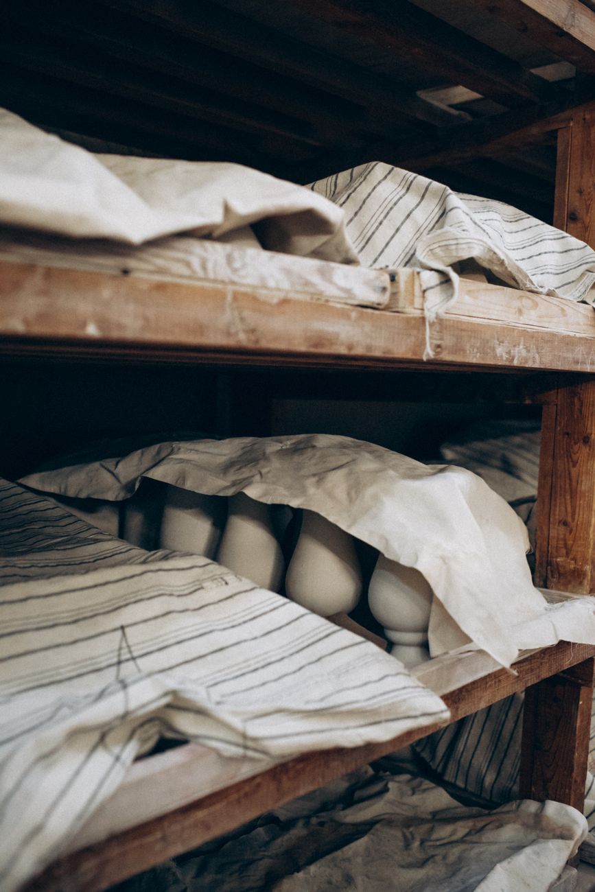 Rustic image of ceramic pots drying under cloth on wooden shelves in a pottery studio.