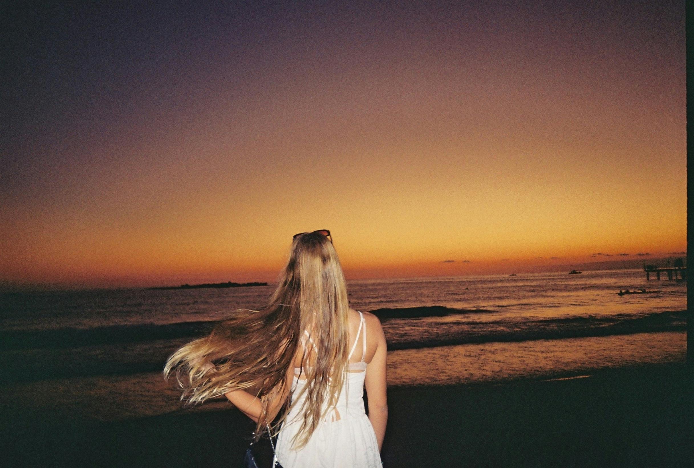 Back view of a young woman with long hair admiring a vibrant ocean sunset.