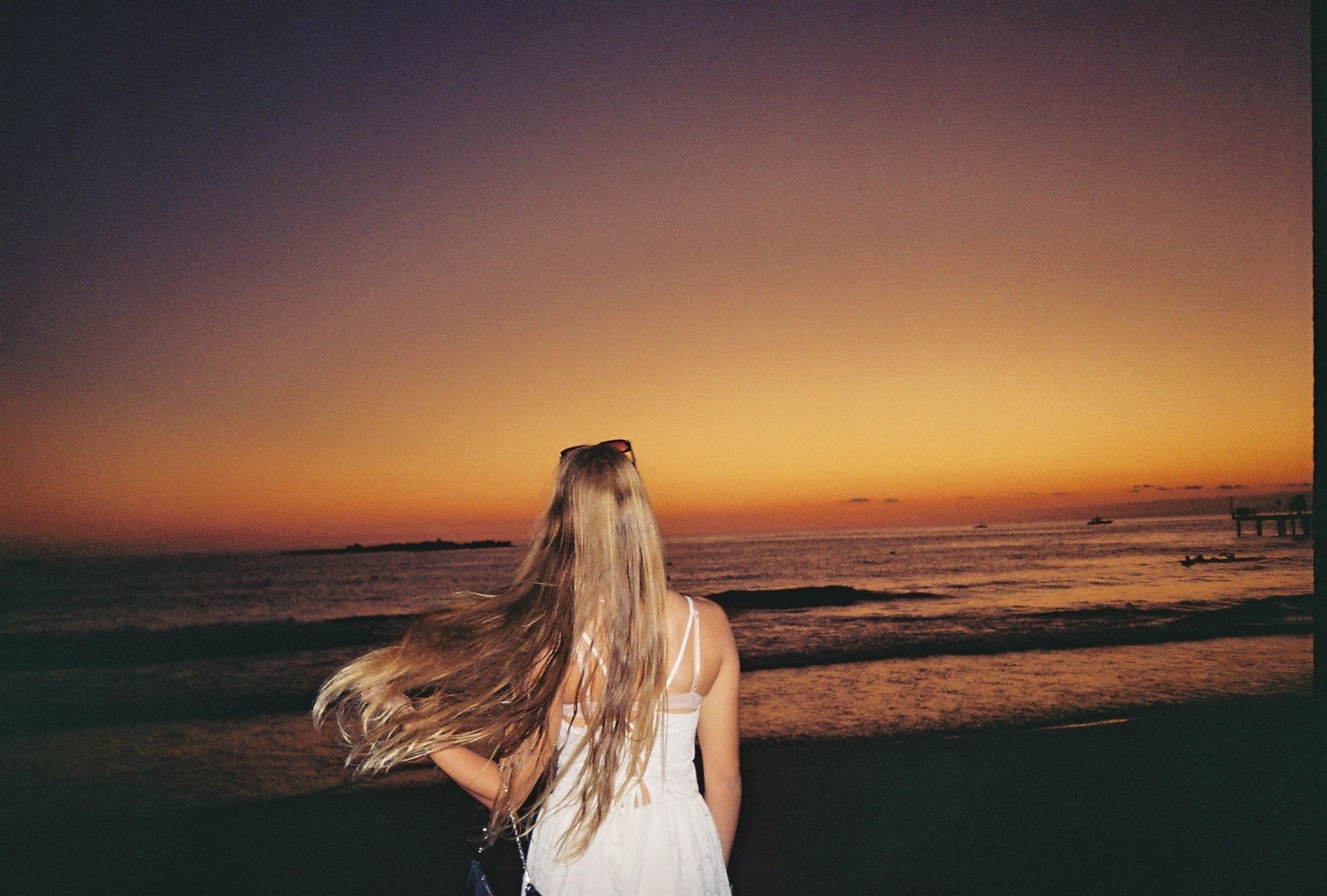 Gratis Vista posteriore di una giovane donna con i capelli lunghi che ammira un vivace tramonto sull'oceano. Foto a disposizione