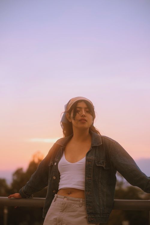 Free A young woman in denim jacket and white top enjoying a serene sunset outdoors, showcasing a peaceful and casual vibe. Stock Photo