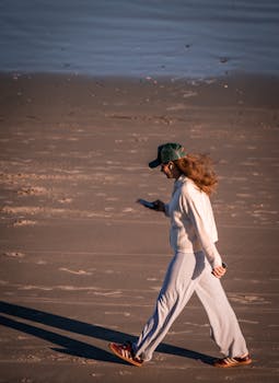 A woman strolls along a sandy beach while focused on her smartphone.